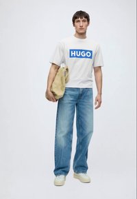 Jeune homme portant un t-shirt blanc "HUGO", jean bleu, baskets blanches, tenant un sac beige à côté, debout devant un fond blanc uni.