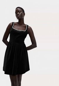 Schwarzes A-Linien-Kleid mit eckigem Ausschnitt, verziert mit einer floralen Applikation entlang des oberen Bereichs, aus weichem Stoff mit strukturierter Oberfläche.