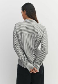 Woman with long dark hair wearing a grey and white striped shirt, standing with hands clasped behind her back against a plain background.