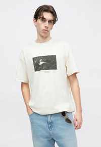 Νέος άνδρας που φοράει κρεμ μπλουζάκι με μαύρο γραφικό διάστημα και κείμενο, ανοιχτόχρωμο μπλε τζιν με γυαλιά ηλίου μπαλωμένα στην τσέπη.