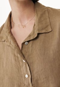 Camicia di lino marrone con colletto, caratterizzata da bottoni bianchi. Una delicata collana d'oro con pendente a forma di mezzaluna è ben visibile.