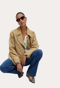 Semišová motorkářská bunda v hnědé barvě s několika zipovými kapsami, nošená přes bílý top. K tomu modré rozšířené džíny a boty s potiskem hada dotvářejí vzhled.