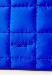 Tissu en nylon bleu matelassé, avec un motif cousu de carrés. Un texte brodé indique "ARKK COPENHAGUE EST. 2015" en bleu.