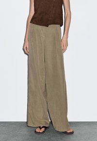 Licht beige wijde broek gemaakt van zacht materiaal, met zijzakken en een ontspannen pasvorm, gecombineerd met bruine sandalen en een mouwloos topje.