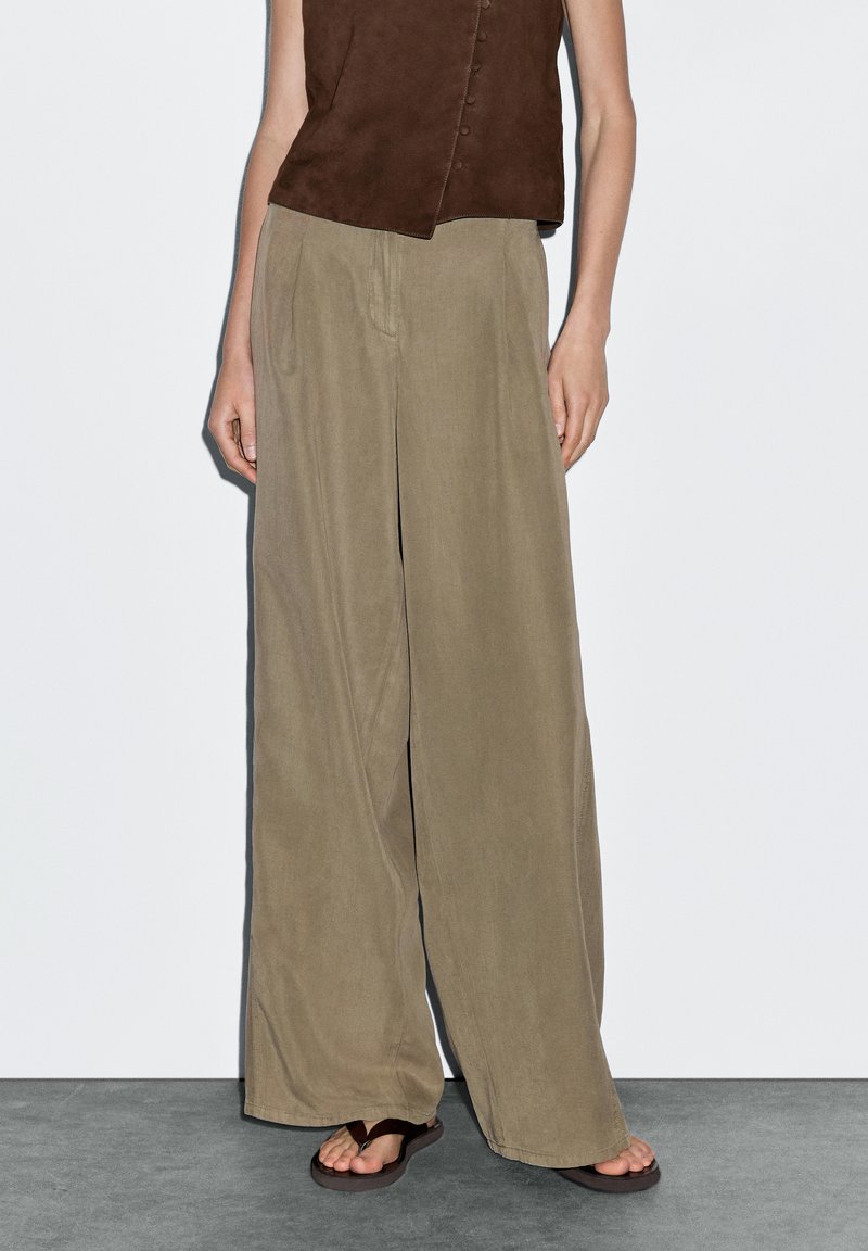 Licht beige wijde broek gemaakt van zacht materiaal, met zijzakken en een ontspannen pasvorm, gecombineerd met bruine sandalen en een mouwloos topje.