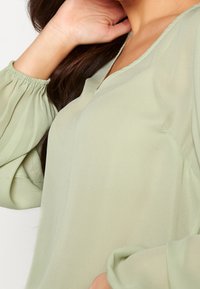 Hellgrüne Bluse mit langen, gerafften Ärmeln und V-Ausschnitt. Hergestellt aus weichem, fließendem Stoff, der eine glatte Textur und dezenten Glanz aufweist.