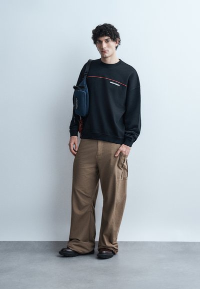 Jonge man met krullend haar, gekleed in een zwart sweatshirt, bruine wijde broekspijpen, zwarte schoenen en een navy crossbodytas, staand tegen een effen muur.