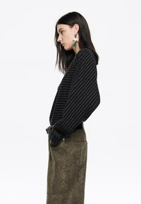 Frau in navy- und olivgestreiftem Pullover und olivfarbener, strukturierter Rock, seitlich stehend mit den Händen in den Taschen vor einem weißen Hintergrund.