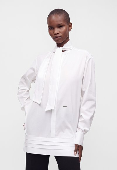 Femme debout aux cheveux courts portant un chemisier blanc à manches longues avec un nœud au col et un ourlet plissé, associé à un pantalon noir sur un fond uni.