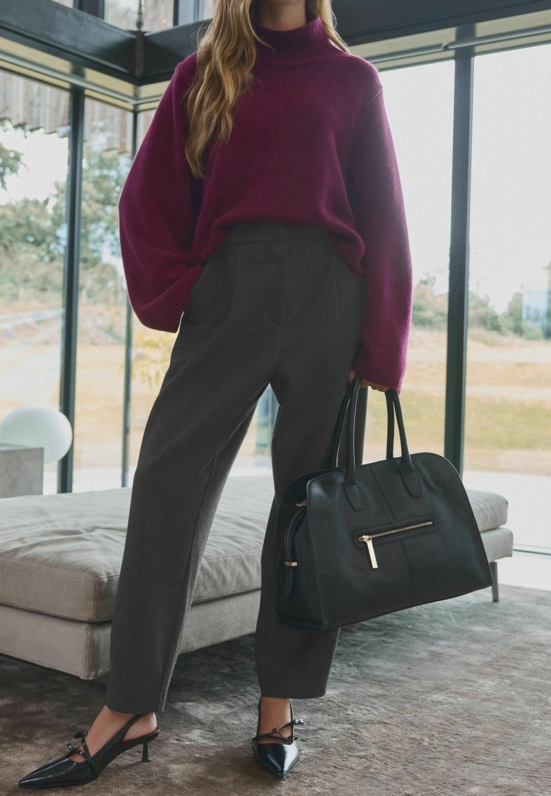 Femme portant un pull violet, un pantalon large foncé, des talons noirs pointus, tenant un grand sac à main noir à l'intérieur près de grandes fenêtres.