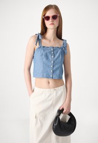 Denim crop top su raukšlių kraštais, mygtukų užsegimu ir reguliuojamomis raištelėmis. Derinama su smėlio spalvos sijonu ir juoda mazgų rankine.