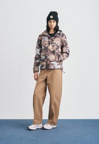 Φλις πουλόβερ σε καφέ και μπεζ με αφηρημένο σχέδιο, με τσέπη με φερμουάρ μπροστά• συνδυασμένο με παντελόνι φαρδιάς γραμμής σε ανοιχτό καφέ και λευκά αθλητικά παπούτσια.