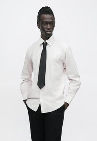 Muž s opletenými vlasmi, oblečený v svetlej pruhovanej košeli na gombíky, čiernej kravate a čiernych nohaviciach, stojaci s rukami v kapse na bielom pozadí.