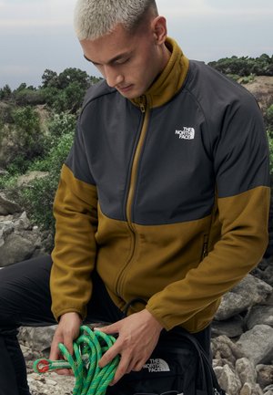 Giacca in fleece di colore grigio scuro e giallo senape con zip, colletto alto e logo The North Face. Modello che tiene una corda da arrampicata verde.