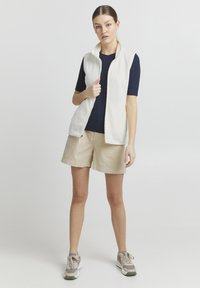 Weißes ärmelloses Oberteil über einem marineblauen gerippten Top, kombiniert mit beigen Shorts. Das Modell trägt leichte Sneakers mit grauen Akzenten. Einfarbiger Hintergrund.