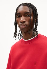 Felpa rossa con scollatura tonda, polsini a costine e un sottile dettaglio con logo. Tessuto liscio, vista frontale del modello con capelli intrecciati.