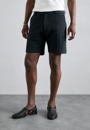 Dunkelgrüne Shorts aus strukturiertem Stoff, mit einer vorderen Falte und Seitentaschen. Kombiniert mit schwarzen Slippern, die ausgeschnittene Akzente zeigen.