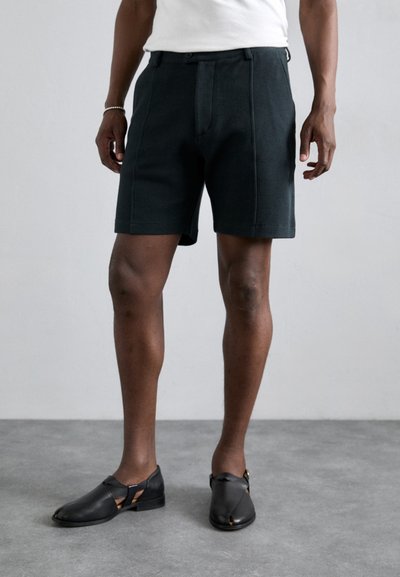 Pantaloni corti verde scuro in tessuto testurizzato, con una piega frontale e tasche laterali. Abbinati a scarpe nere slip-on con dettagli a taglio.