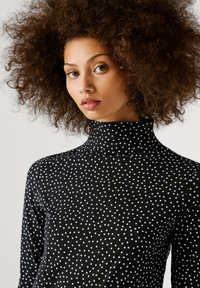 Hölgy, kiterjedt göndör hajjal, fekete garbóban, amelyet kis fehér pöttyök borítanak, enyhén oldalra nézve, egyszínű háttér előtt.