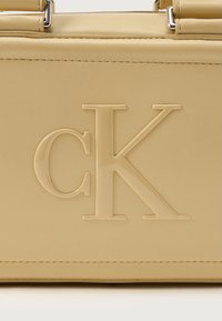 Sac à main Calvin Klein en cuir beige texturé avec logo 'CK' en relief, matériel en argent et lignes épurées avec un minimum de couture.
