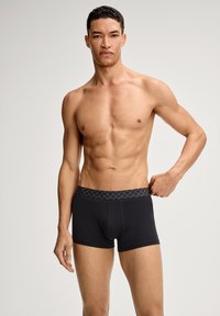 Musta bokserialushousut, joiden joustavassa vyötärönauhassa on pyöreä kuvio. Kangas näyttää pehmeältä ja vartalonmyötäiseltä.