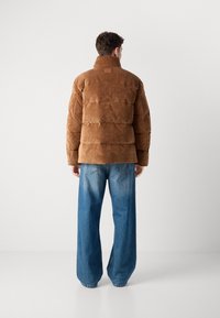 Braune Corduroy-Puffjacke mit hohem Kragen, gestepptem Design und einem Lederpatch am Hals, kombiniert mit weit geschnittenen blauen Jeans.