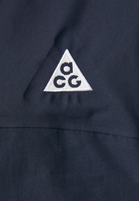 Tamno plavi materijal s bijelim izvezenim trokutastim logotipom s malim slovima "a", "c" i "g". Glatka tekstura s suptilnim sjajem.