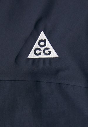 Ciemnoniebieska tkanina z białym, haftowanym trójkątnym logo z małymi literami "a", "c" i "g". Gładka tekstura z subtelnym połyskiem.