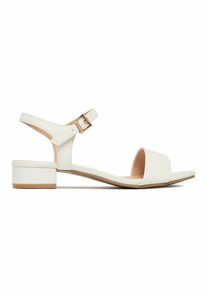 White low-heeled sandal with open toe, wide front strap, ankle strap with gold buckle, and a beige sole against a white background.