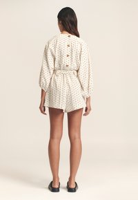 Combinaison à pois en tissu crème, avec des manches longues, une taille ceinturée et un dos boutonné. Coupe courte avec des détails texturés.