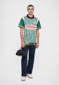 Green and white striped short-sleeve shirt with collar, featuring a logo. Dark jeans, white shoes, and a small black handbag with handles.