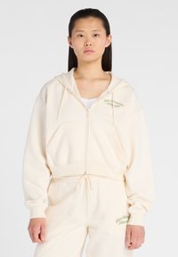 Sweat à capuche zippé crème avec poches avant, texte brodé en vert clair et coupe décontractée. Assorti avec un pantalon de jogging crème.