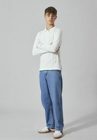 Langärmeliges weißes Poloshirt kombiniert mit hellblauen geraden Jeans und weißen Sneakern mit schwarzen Akzenten, vor einem grauen Hintergrund stehend.