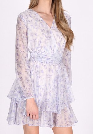 Kobieta ubrana w przezroczystą, długorękawową sukienkę kopertową w jasnofioletowe kwiaty, z warstwową spódnicą i talią podkreśloną paskiem.