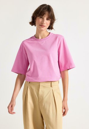 Nuori nainen, jolla on lyhyet ruskeat hiukset, pukeutunut vaaleanpunaiseen ylisuurikokoiseen T-paitaan ja beigenvärisiin korkeavyötäröisiin laskostettuihin housuihin, seisomassa yksinkertaista taustaa vasten.