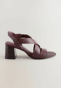 Burgundy leather sandal with a block heel. Features crisscross straps and an open back. Smooth texture and rounded toe design.