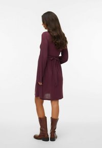 Mavri obleka z dolgimi rokavi in prilagojenim pasom, razširjenim krilom in dolžino do kolen, v kombinaciji z rjavimi škornji do kolen, gladka tekstura.