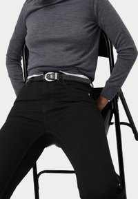 Černé džíny s úzkým střihem a poutky na opasek, doplněné šedým topem s dlouhým rukávem. Lesklý černý opasek se stříbrnou sponou zvýrazňuje pas.