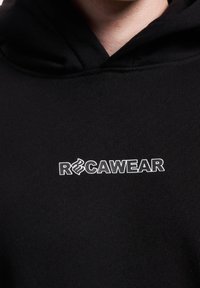 Sort hættetrøje lavet af blødt stof, med et hvidt logo der står "RCAWEAR" med et stiliseret bogstavdesign, fremtrædende nær halsudskæringen.