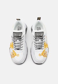 Bílé tenisky s černými akcenty, s jemnou žlutou květinovou výšivkou, texturovanými tkaničkami a výrazným logem na jazyku.