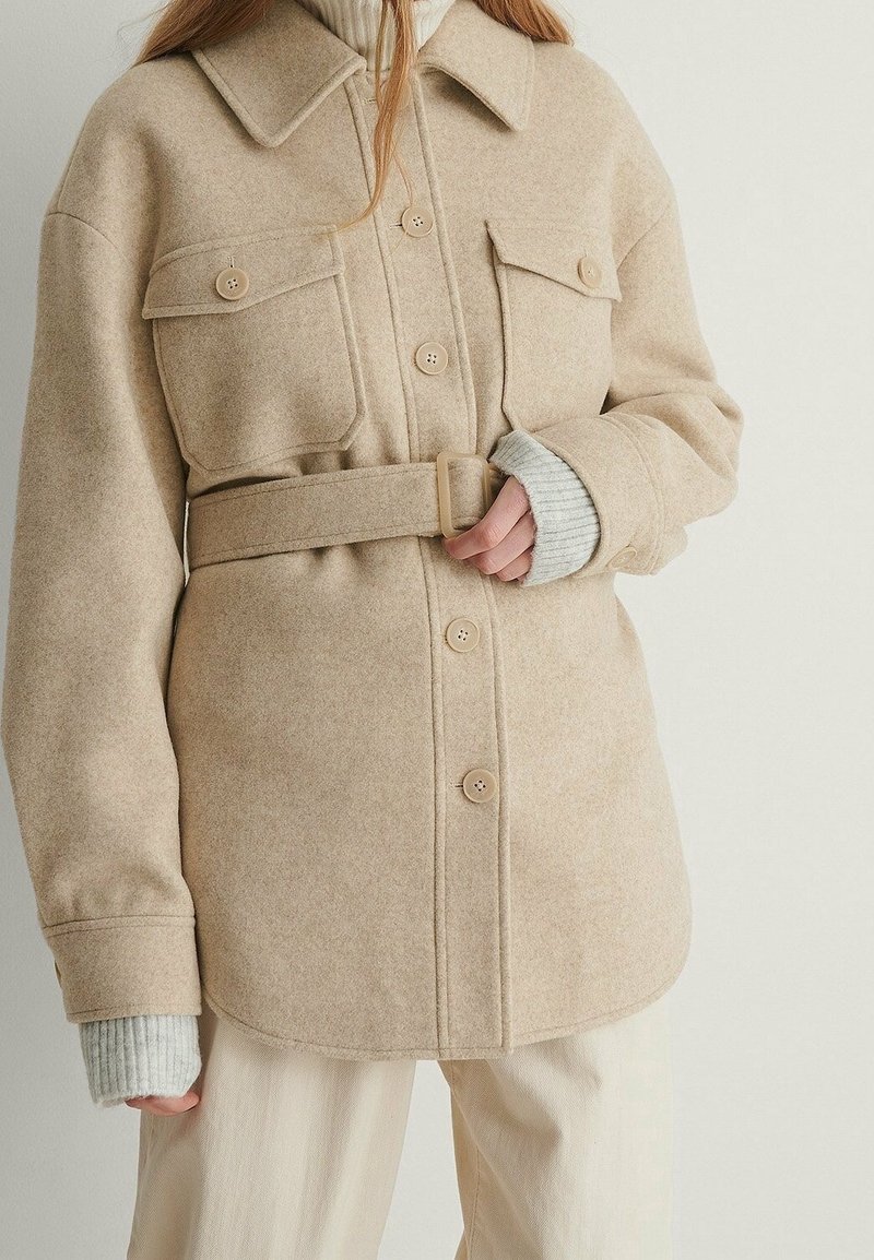 Kvinde iført beige uldjakke med bælte, store forlommer og knapper, lagdelt over en grå ribstrikket trøje og cremefarvede bukser.