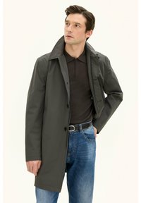 Mörkgrå vattentät kappa med hög krage, knappstängning och sidfickor, i kombination med en brun polo och blå jeans. Slät textur.