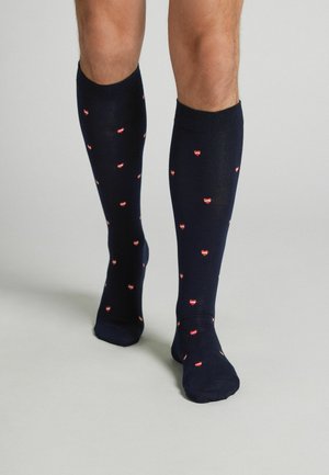 Beine mit marineblauen knielangen Socken, die mit kleinen roten Herzen mit dem Wort „LOVE“ auf hellgrauem Hintergrund gemustert sind.
