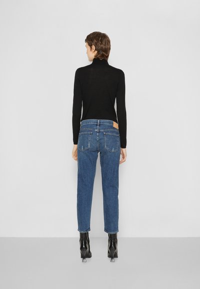Schwarzes Langarmoberteil, kombiniert mit hochgeschnittenen blauen Jeans aus Denim. Die Jeans haben eine helle Waschung und zwei hintere Taschen, vollendet mit schwarzen Stiefeln mit Absatz.