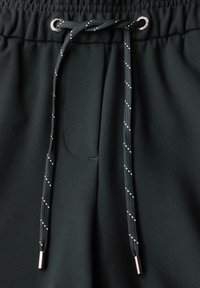Un pantalon de sport vert foncé présente un cordon de serrage avec des embouts en argent et un motif texturé. Tissu lisse avec un détail de poche latérale.