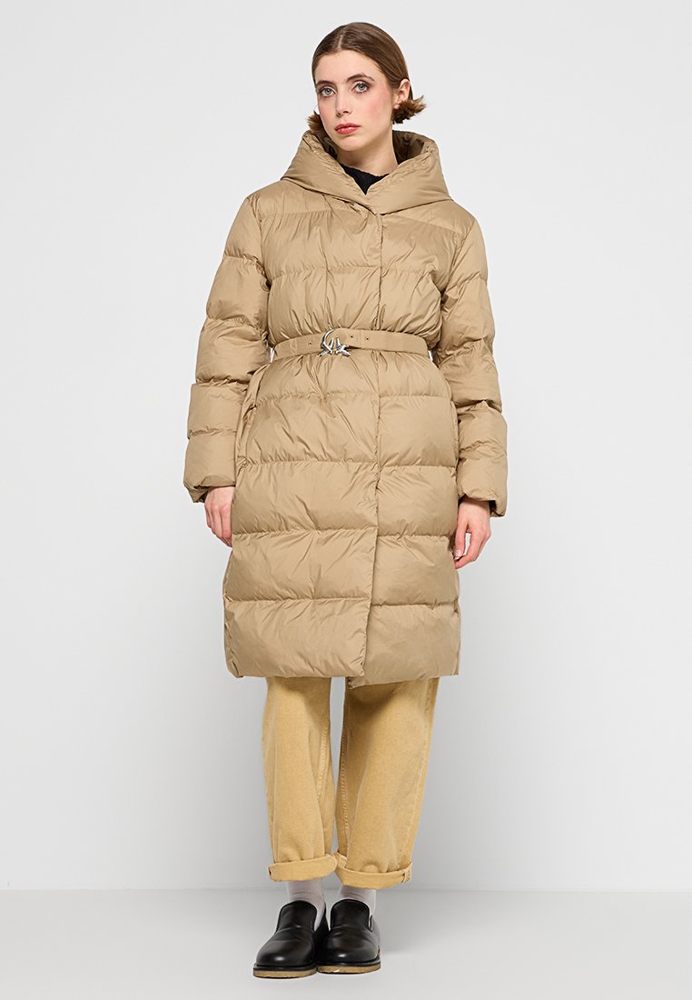 Cappotto puffer beige con cintura arricciata e fibbia argentata, dotato di colletto alto, maniche lunghe e texture trapuntata. Indossato sopra pantaloni di colore chiaro.