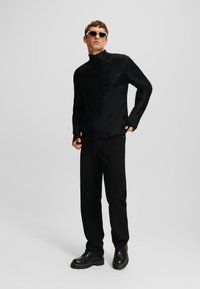 Sort strikket turtleneck sweater med struktureret mønster, parret med sorte lige ben bukser og sorte snørebåndssko; modellen bærer solbriller.