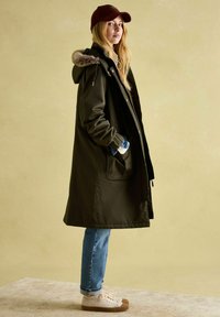 Long dark green parka with a fur-lined hood, side pockets, and button closure, paired with light blue jeans and beige sneakers. Burgundy cap worn.