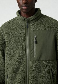 Giacca in pile verde oliva con esterno strutturato, chiusura con zip, collo alla coreana e tasche frontali. Presenta dettagli in tessuto liscio e accessori in metallo.