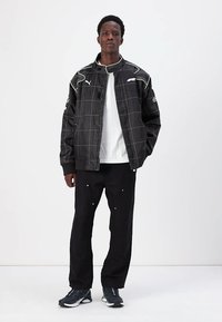 Black bomber jacket with grid pattern, ribbed cuffs, and PUMA logo. Worn over a white shirt, paired with black pants and gray sneakers.